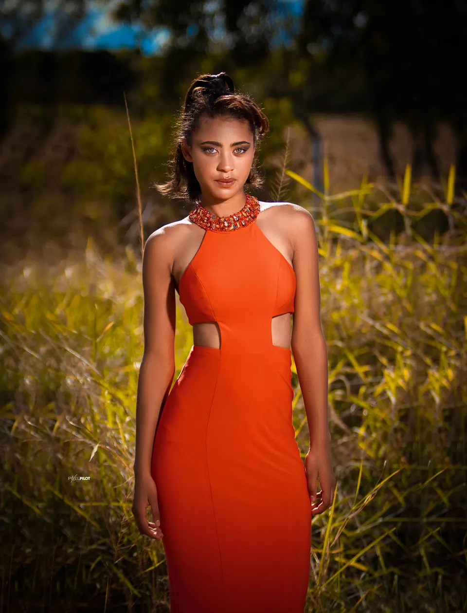 Portrait of a woman in an orange halter gown standing in a golden grass field at sunset